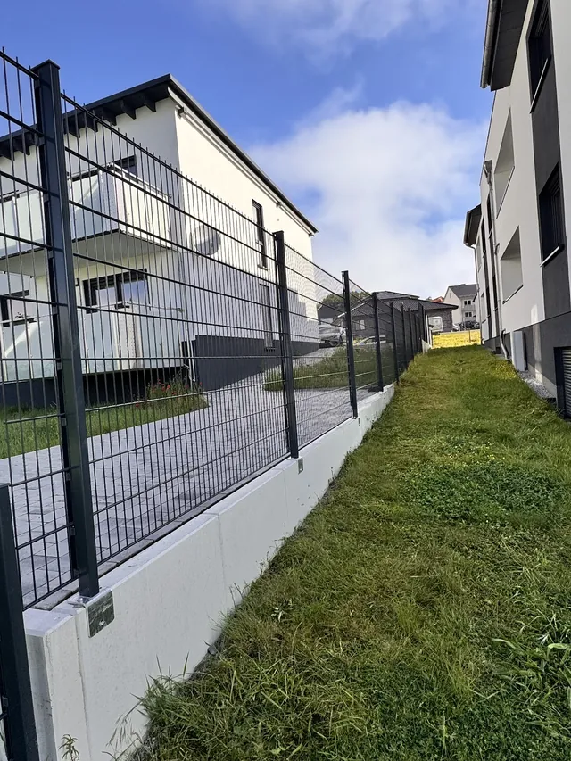 Doppelstabmattenzaun auf einem weißen Betonsockel entlang eines Mehrfamilienhauses