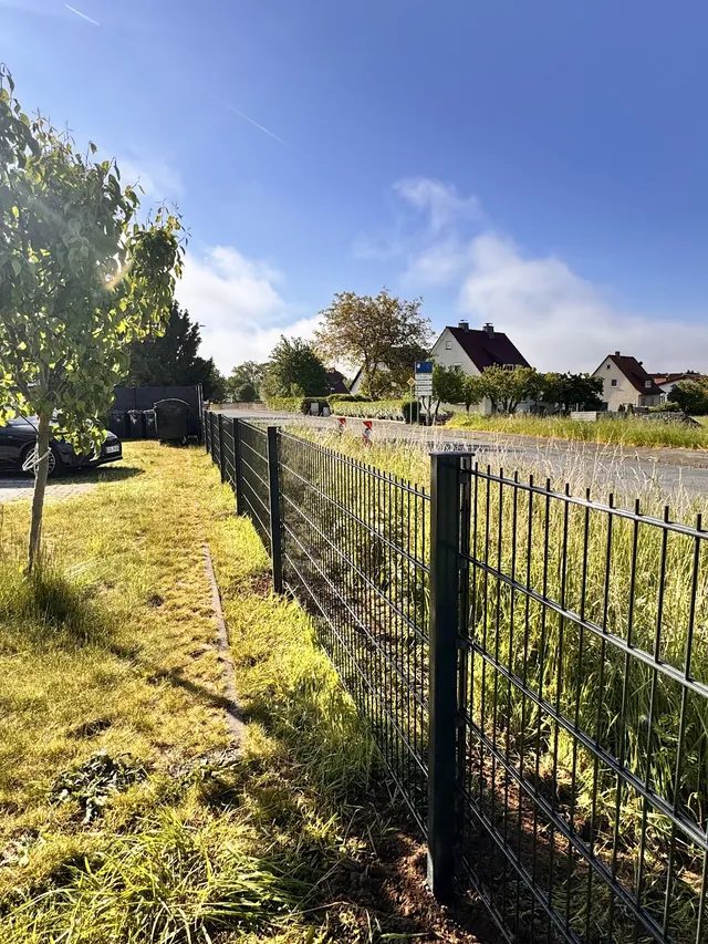 Zaun entlang einer Wiese am Feldweg, im Hintergrund Wohnhäuser