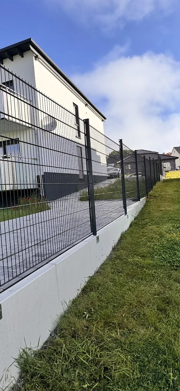 Doppelstabmattenzaun auf weißem Betonsockel entlang eines Mehrfamilienhauses